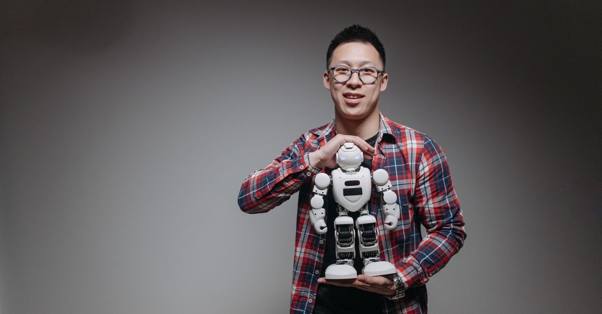Asian man with eyeglasses holding a toy robot in a studio with a gray background.