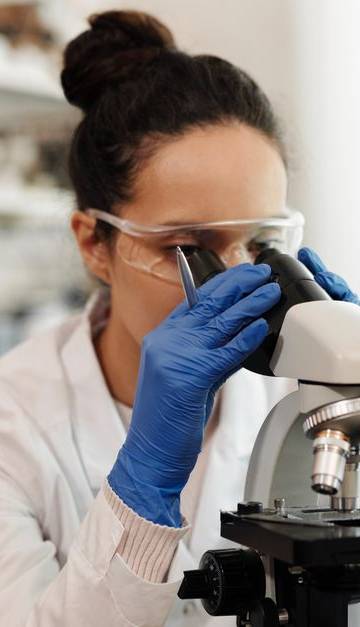 A female scientist in protective gear examines samples through a microscope in a laboratory setting.