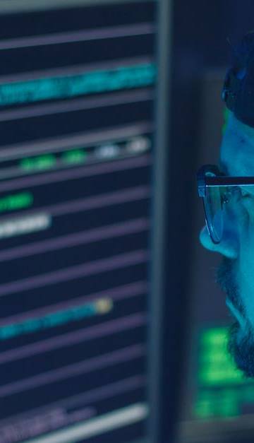A cybersecurity expert inspecting lines of code on multiple monitors in a dimly lit office.