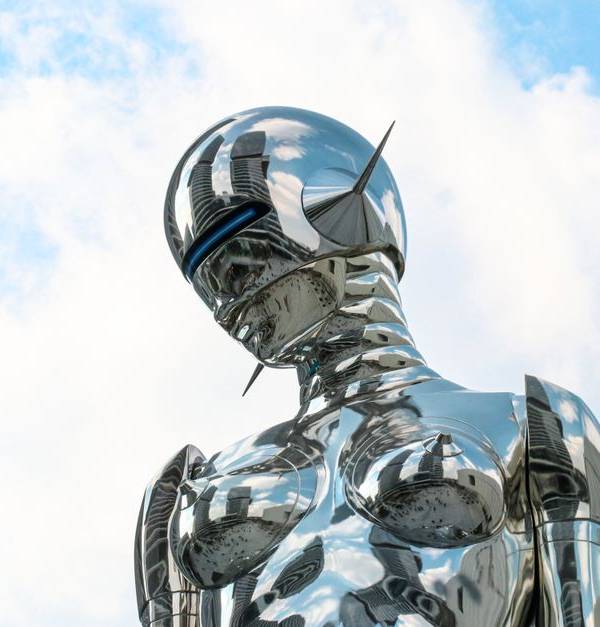 A sleek chrome robot sculpture stands against a bright blue sky background.