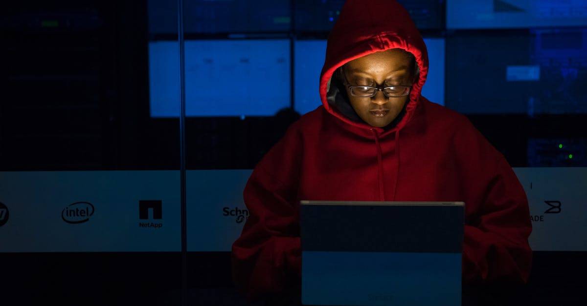 A woman in red hoodie works intensely on her laptop indoors, illuminated by screen light.