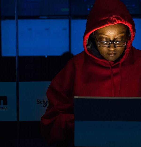A woman in red hoodie works intensely on her laptop indoors, illuminated by screen light.