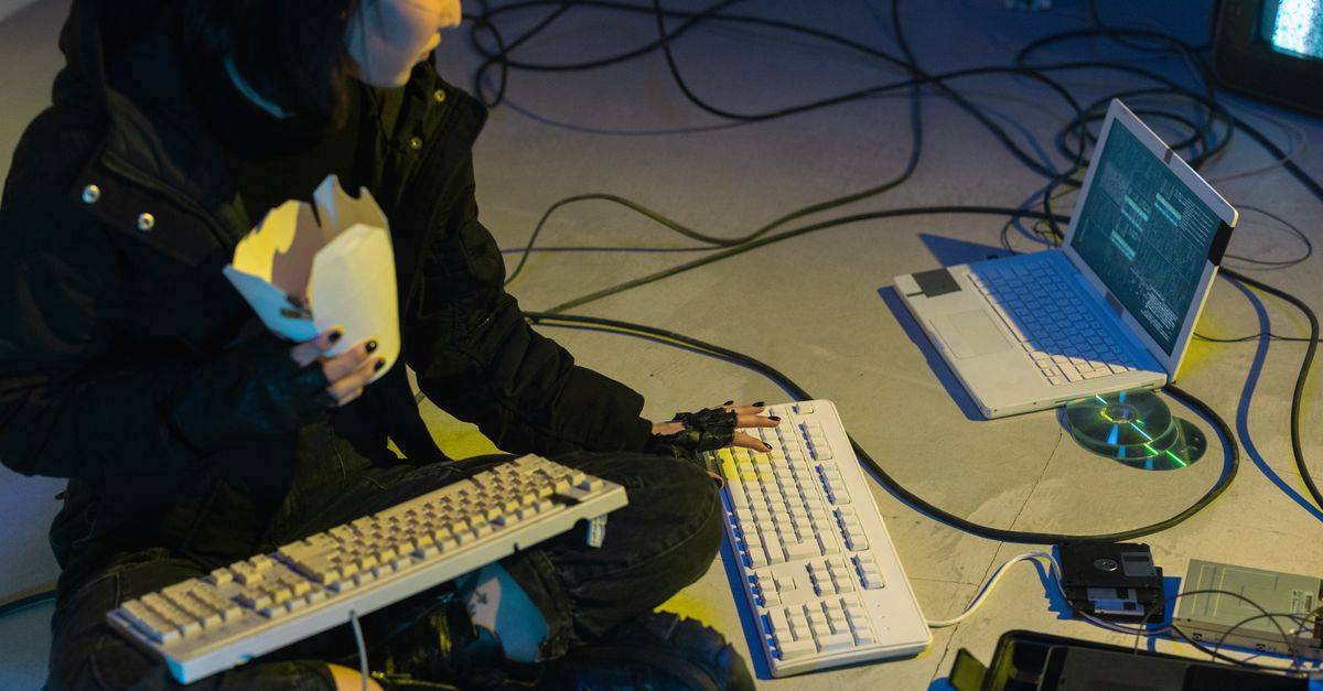 A young woman in a dark room surrounded by computers and cables, eating and typing on keyboards.