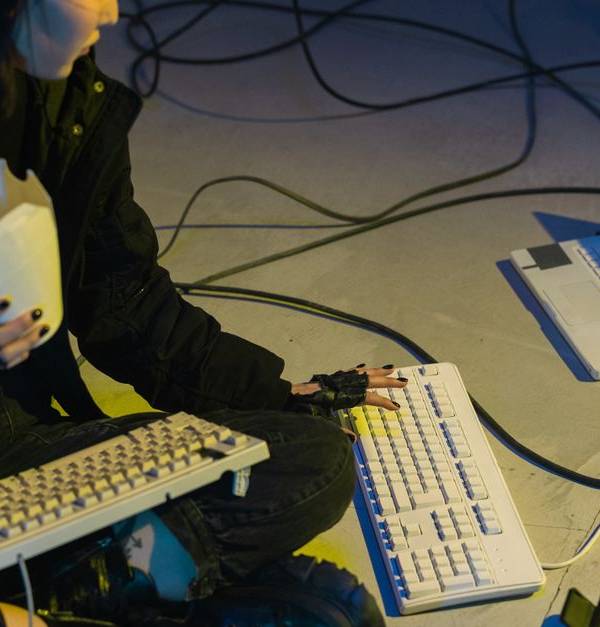 A young woman in a dark room surrounded by computers and cables, eating and typing on keyboards.