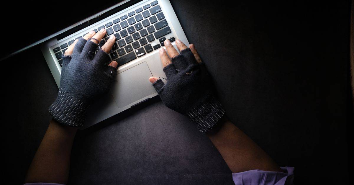 Hacker in fingerless gloves typing on laptop keyboard from above in a dark setting.