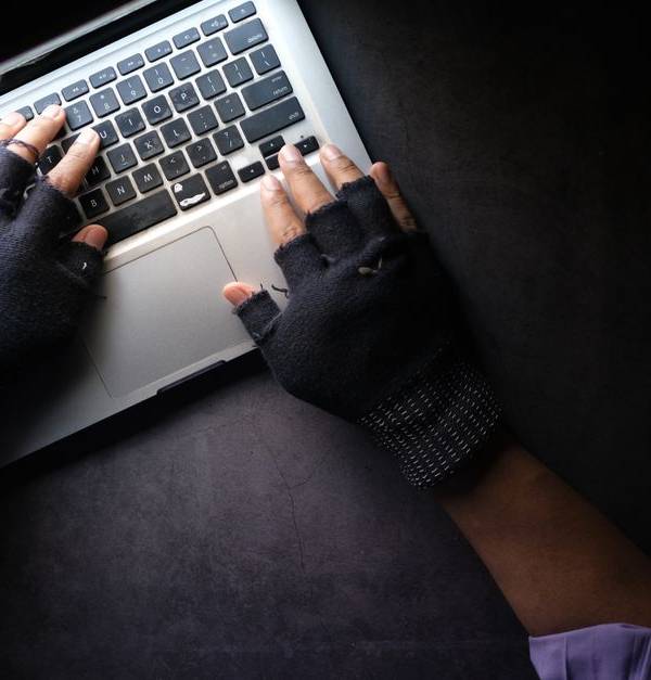Hacker in fingerless gloves typing on laptop keyboard from above in a dark setting.