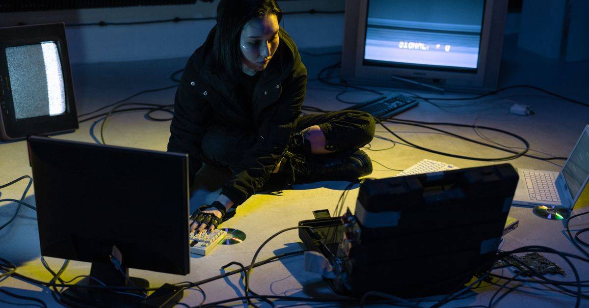 Asian woman working on multiple devices in a tech-heavy environment, illuminated by screen light.