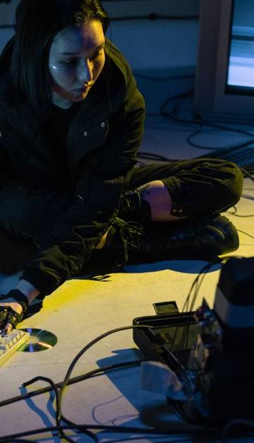Asian woman working on multiple devices in a tech-heavy environment, illuminated by screen light.
