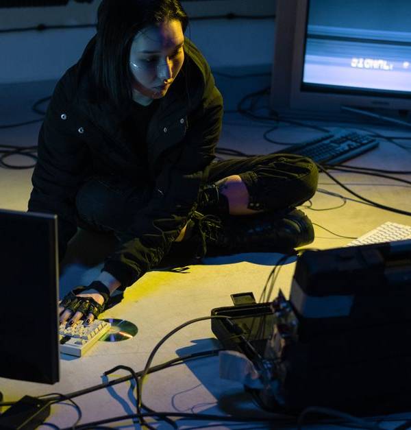 Asian woman working on multiple devices in a tech-heavy environment, illuminated by screen light.