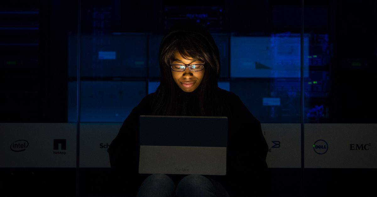 A woman uses her laptop in a dimly lit server room, focusing on technology and work.