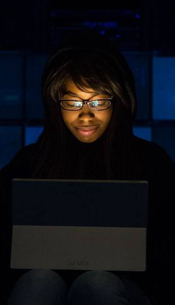 A woman uses her laptop in a dimly lit server room, focusing on technology and work.