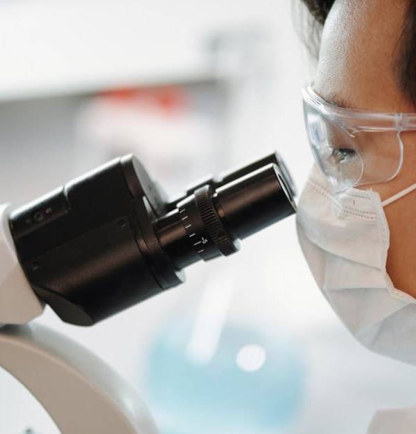 Scientist wearing face mask and goggles examining samples through a microscope in a lab.