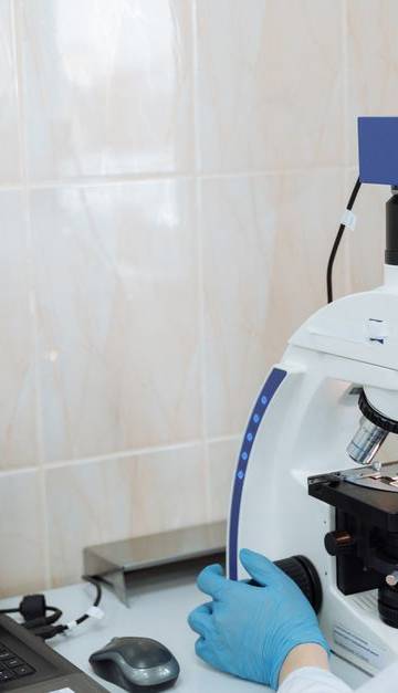 Scientist in lab coat using microscope and laptop in a laboratory setting.