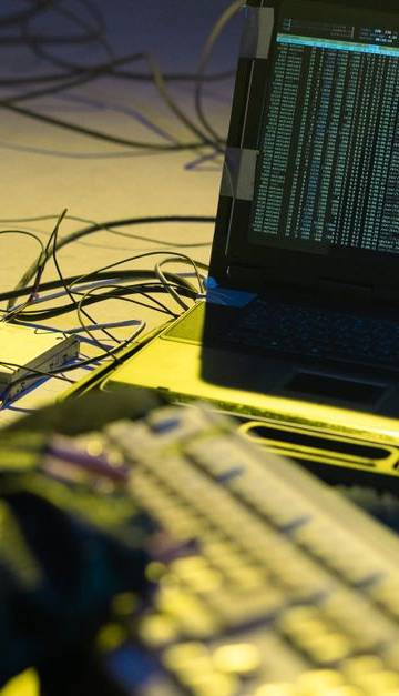 A person typing on a keyboard connected to a laptop showing code, surrounded by wires.