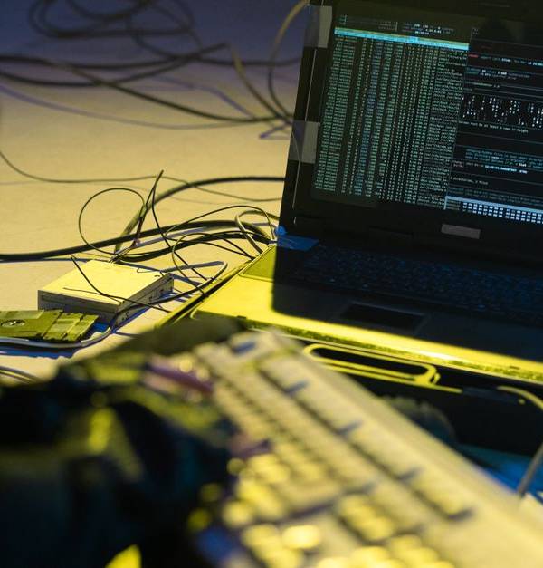 A person typing on a keyboard connected to a laptop showing code, surrounded by wires.
