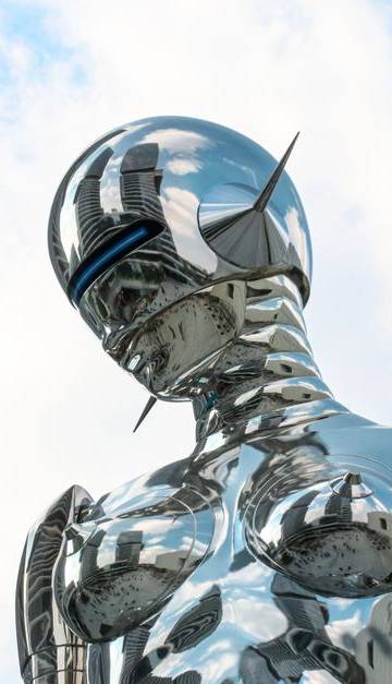 A sleek chrome robot sculpture stands against a bright blue sky background.