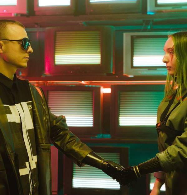 Two individuals with prosthetic arms engage in a handshake amidst a vibrant, retro television backdrop.