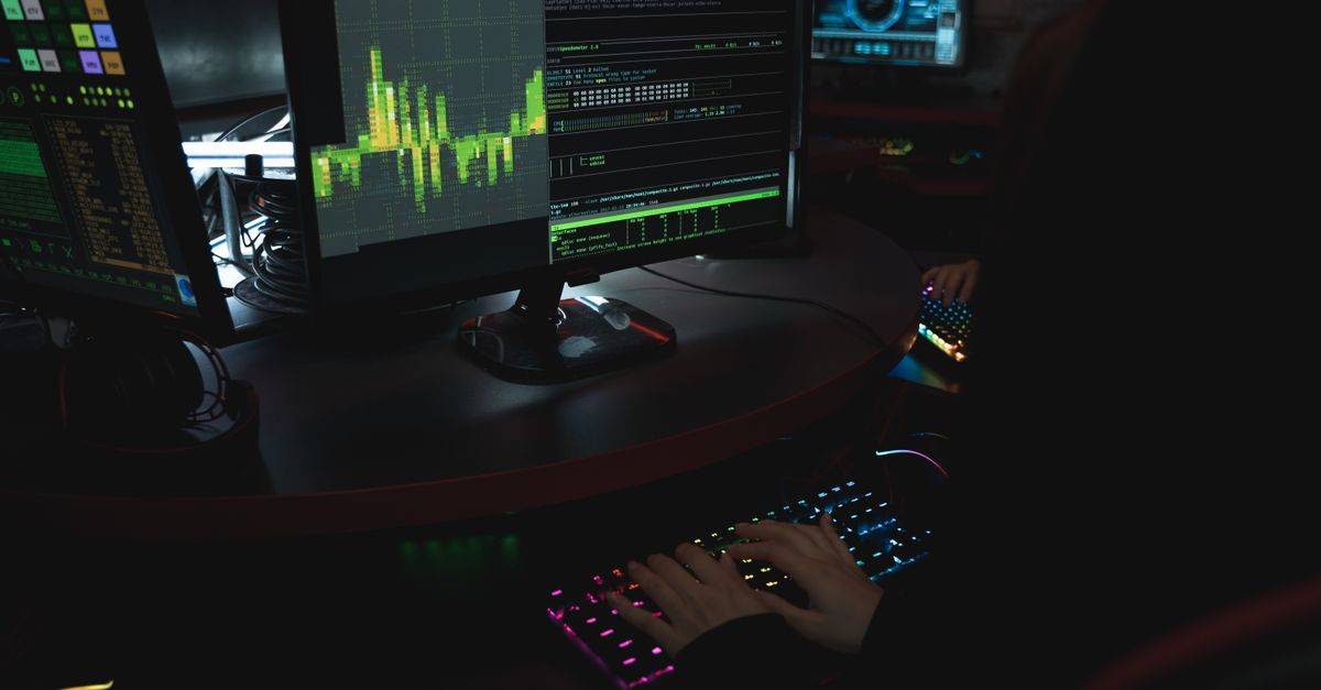 A cybersecurity expert in a dimly lit room is typing on a colorful keyboard with multiple screens displaying data.