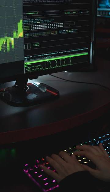 A cybersecurity expert in a dimly lit room is typing on a colorful keyboard with multiple screens displaying data.
