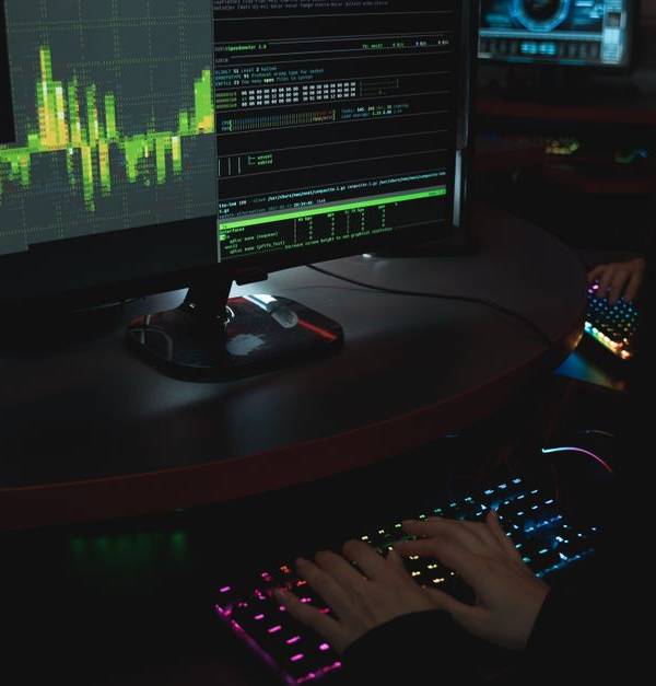 A cybersecurity expert in a dimly lit room is typing on a colorful keyboard with multiple screens displaying data.