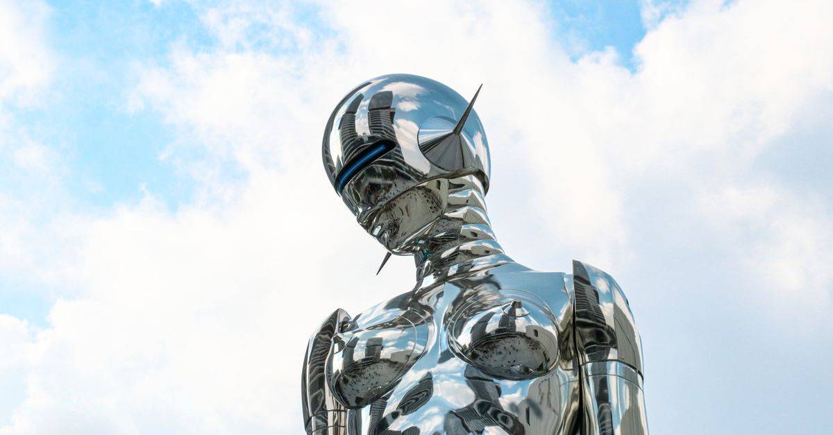 A sleek chrome robot sculpture stands against a bright blue sky background.