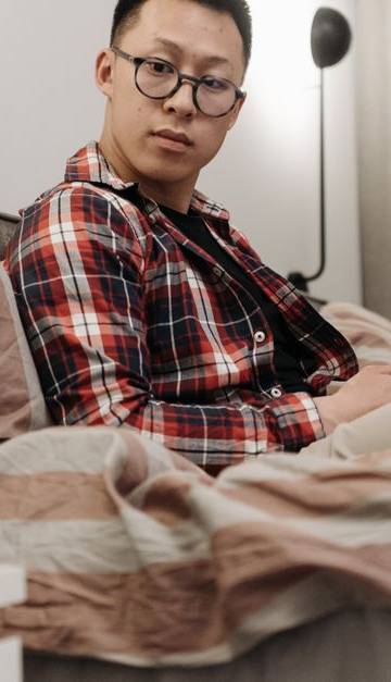 Asian man in plaid shirt sitting on bed, observing a robot toy in a cozy bedroom setting.
