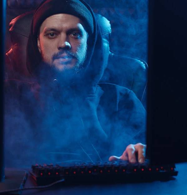 Dramatic scene of an adult man in a hoodie using a computer, surrounded by smoke in a dimly lit room.