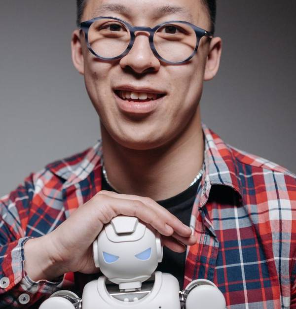 Asian man smiling, holding a small toy robot during a studio shoot. Indoors, with eyeglasses and plaid shirt.