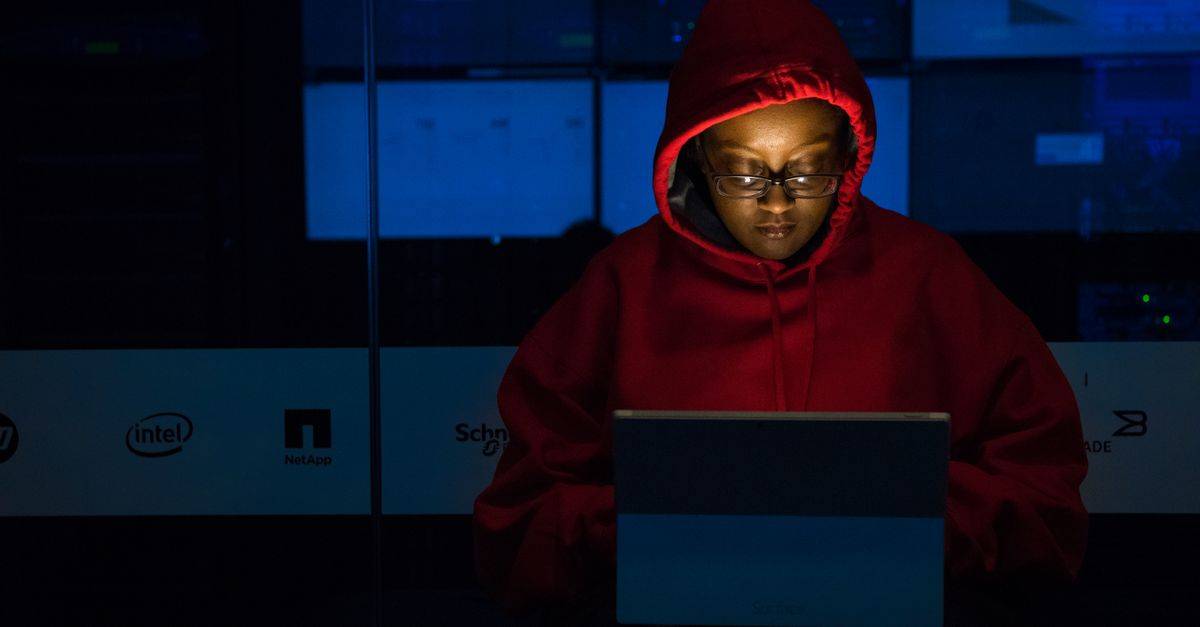A woman in red hoodie works intensely on her laptop indoors, illuminated by screen light.