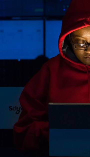 A woman in red hoodie works intensely on her laptop indoors, illuminated by screen light.