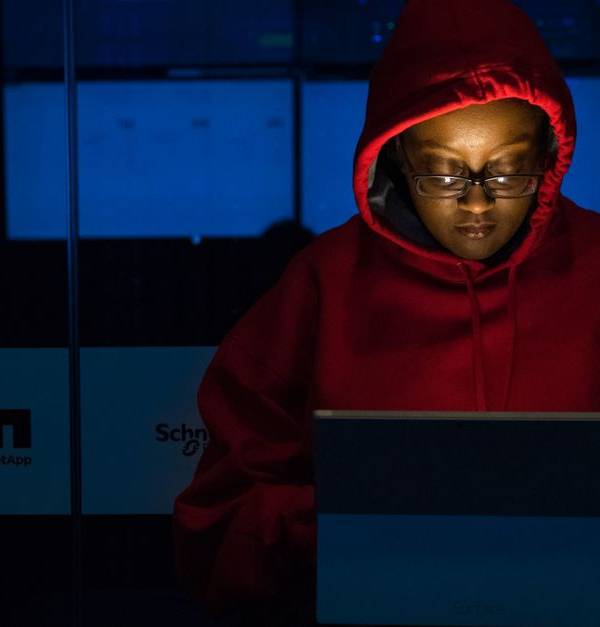 A woman in red hoodie works intensely on her laptop indoors, illuminated by screen light.
