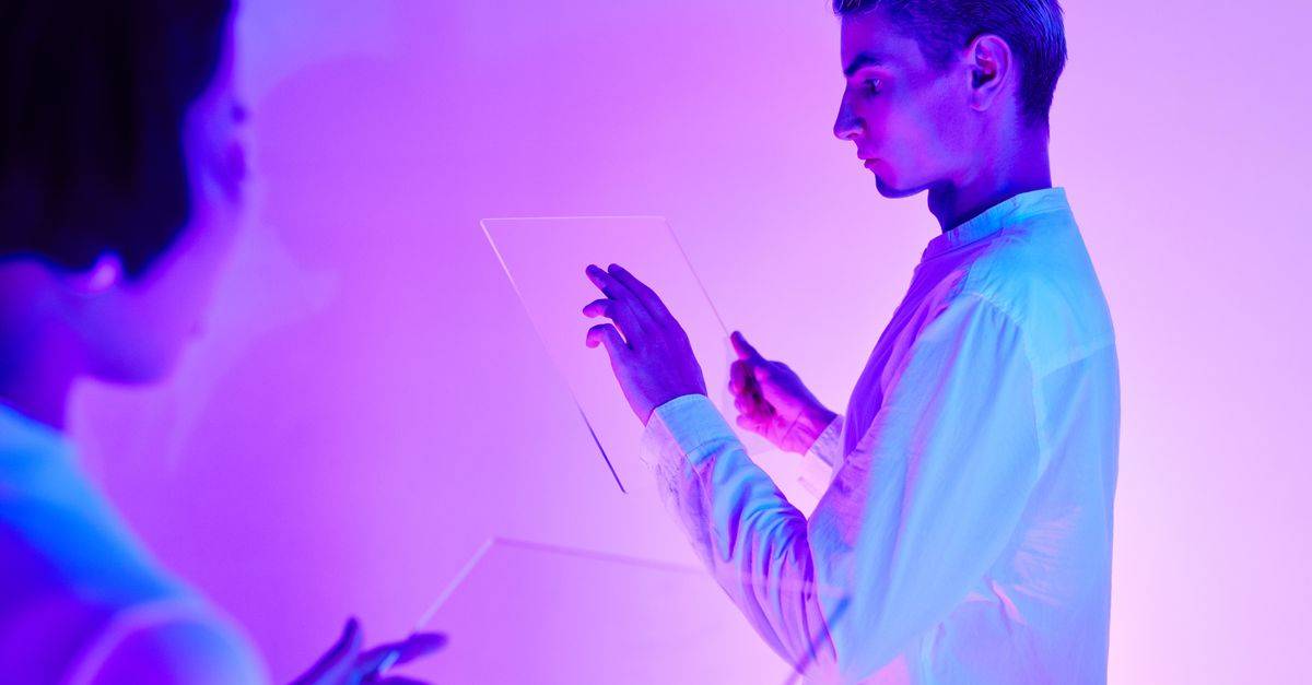 Two individuals engaging with futuristic transparent touch screens in a vibrant purple-lit studio.
