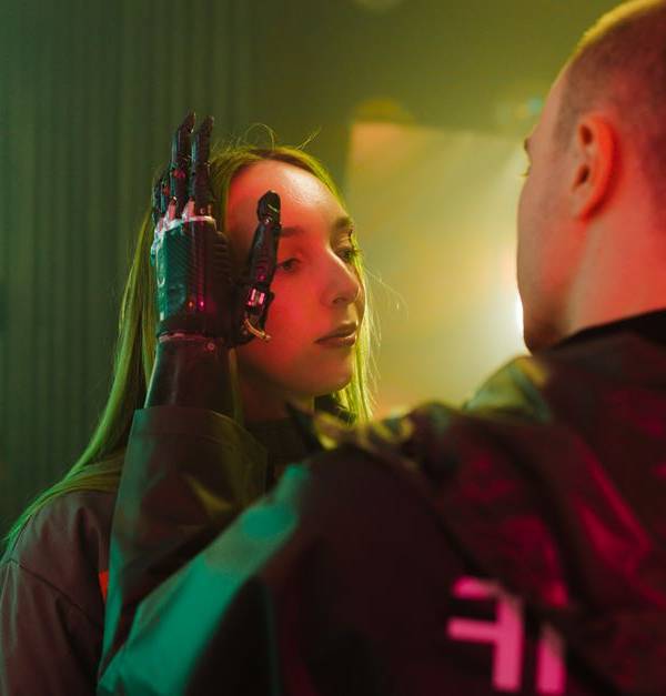 A man with a prosthetic hand gently touches a woman's face in a cyberpunk setting.