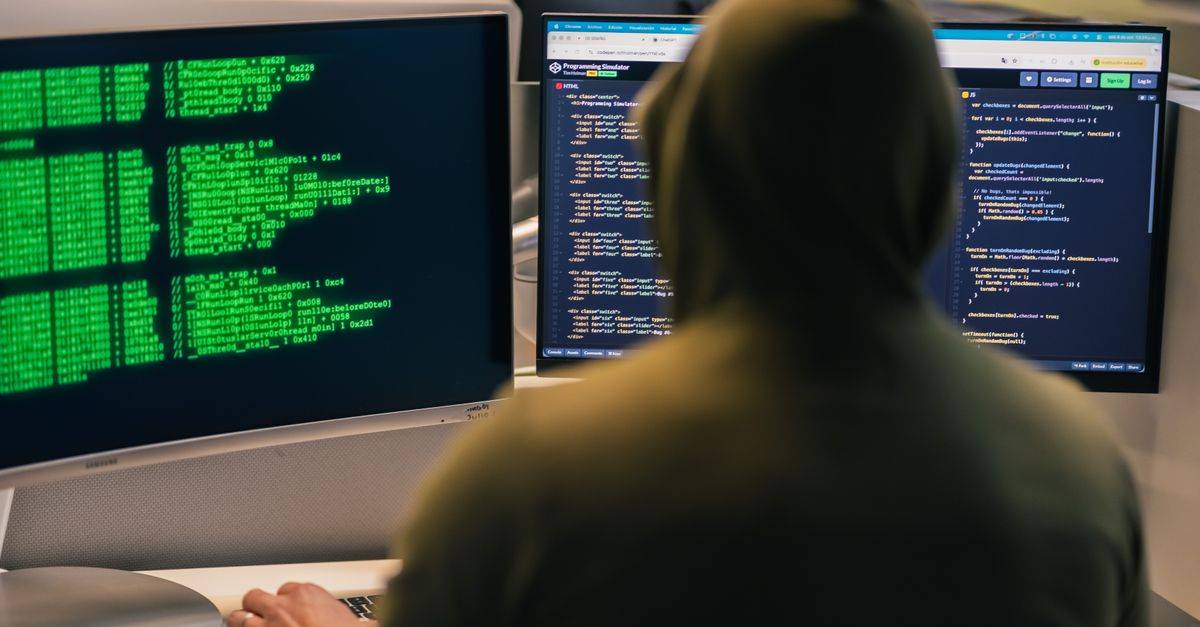 A person in a hoodie coding on dual monitors, depicting cybersecurity and hacking themes.