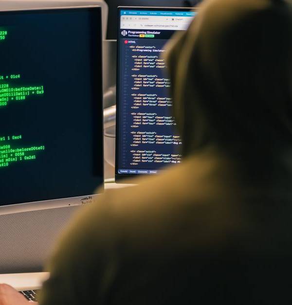 A person in a hoodie coding on dual monitors, depicting cybersecurity and hacking themes.