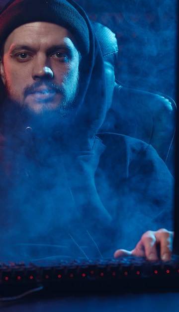 Dramatic scene of an adult man in a hoodie using a computer, surrounded by smoke in a dimly lit room.