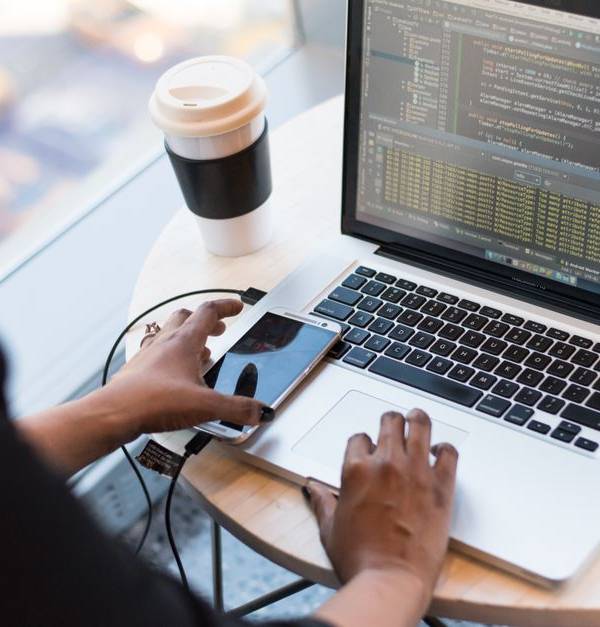 Black woman programming on a laptop with coffee, smartphone, and glasses on a desk in an office.