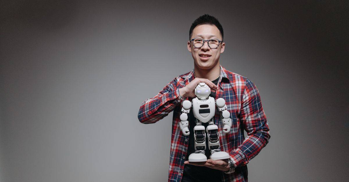 Asian man with eyeglasses holding a toy robot in a studio with a gray background.