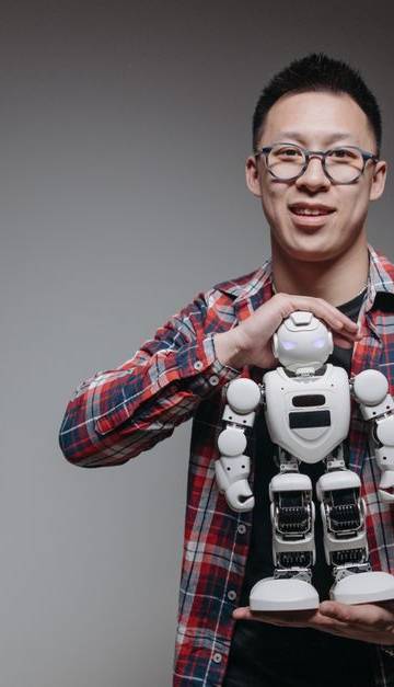 Asian man with eyeglasses holding a toy robot in a studio with a gray background.