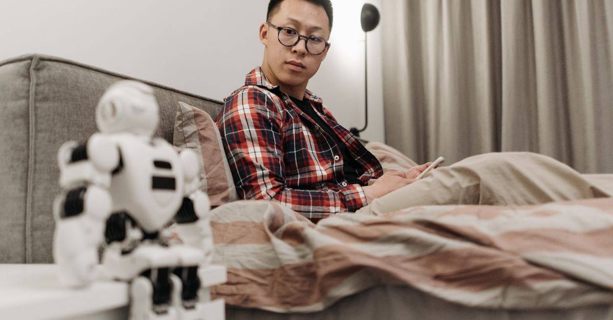 Asian man in plaid shirt sitting on bed, observing a robot toy in a cozy bedroom setting.