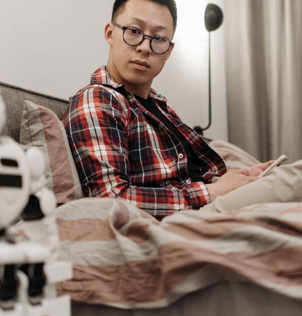 Asian man in plaid shirt sitting on bed, observing a robot toy in a cozy bedroom setting.