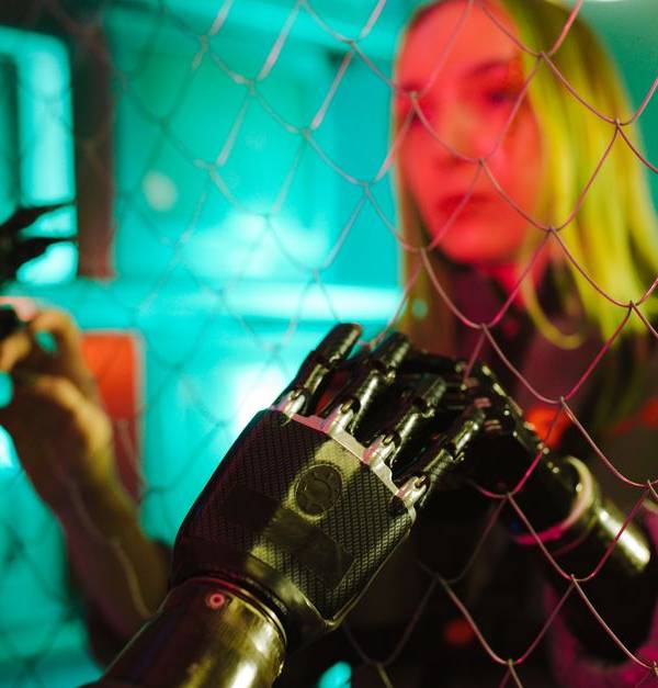 A woman interacts with a robotic hand through a fence under neon lights.