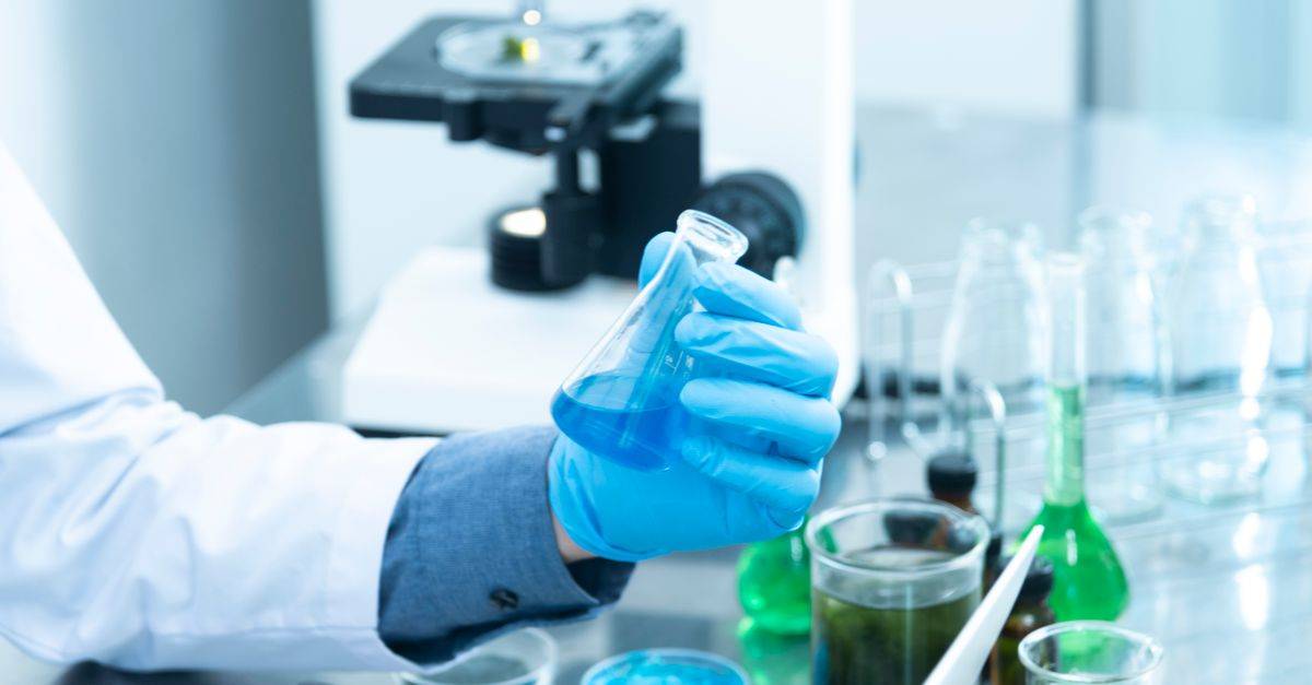 Scientist in gloves analyzing blue liquid in a laboratory setting with microscope and glassware.