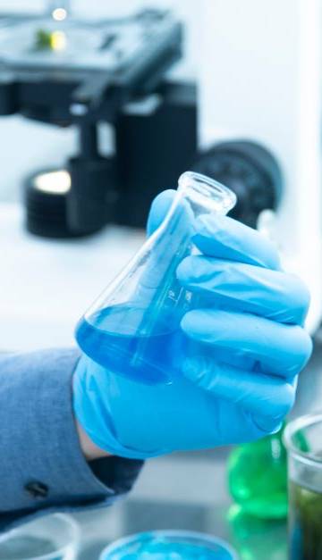 Scientist in gloves analyzing blue liquid in a laboratory setting with microscope and glassware.