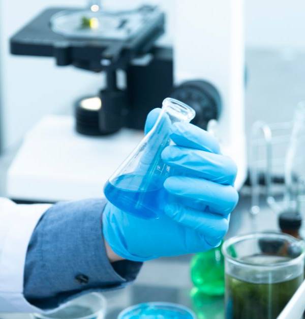 Scientist in gloves analyzing blue liquid in a laboratory setting with microscope and glassware.