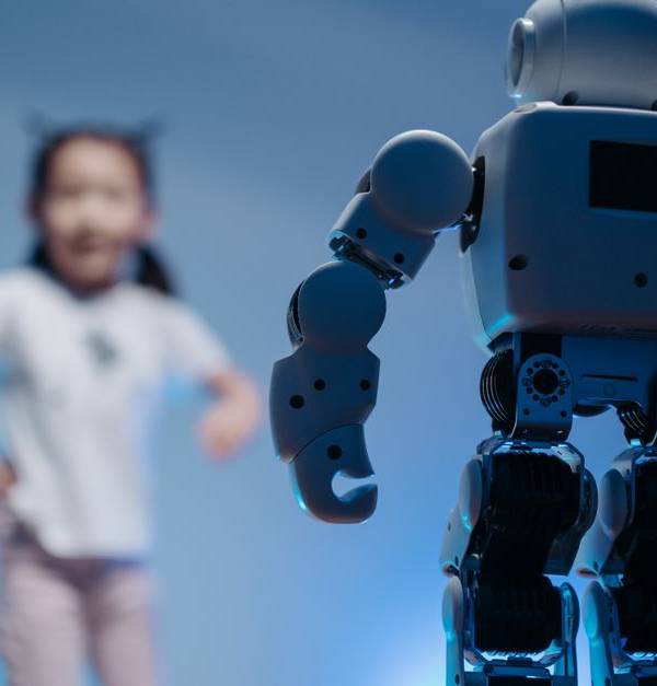 A young girl playfully interacts with a humanoid robot in a futuristic indoor environment featuring soft blue lighting.