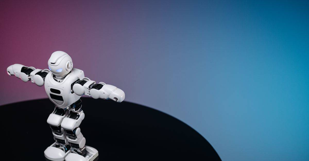 A white humanoid toy robot standing on a reflective black surface in a studio setting with a blue and pink gradient background.